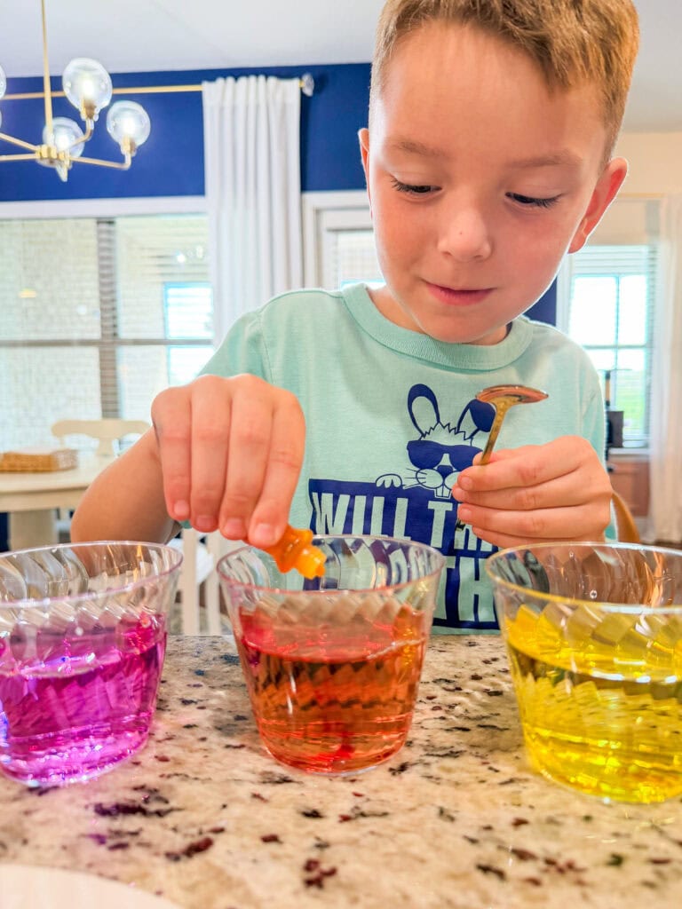 A young boy drops colored liquid into glass cups filled with pink, orange, and yellow water, conducting a science experiment at a kitchen counter.