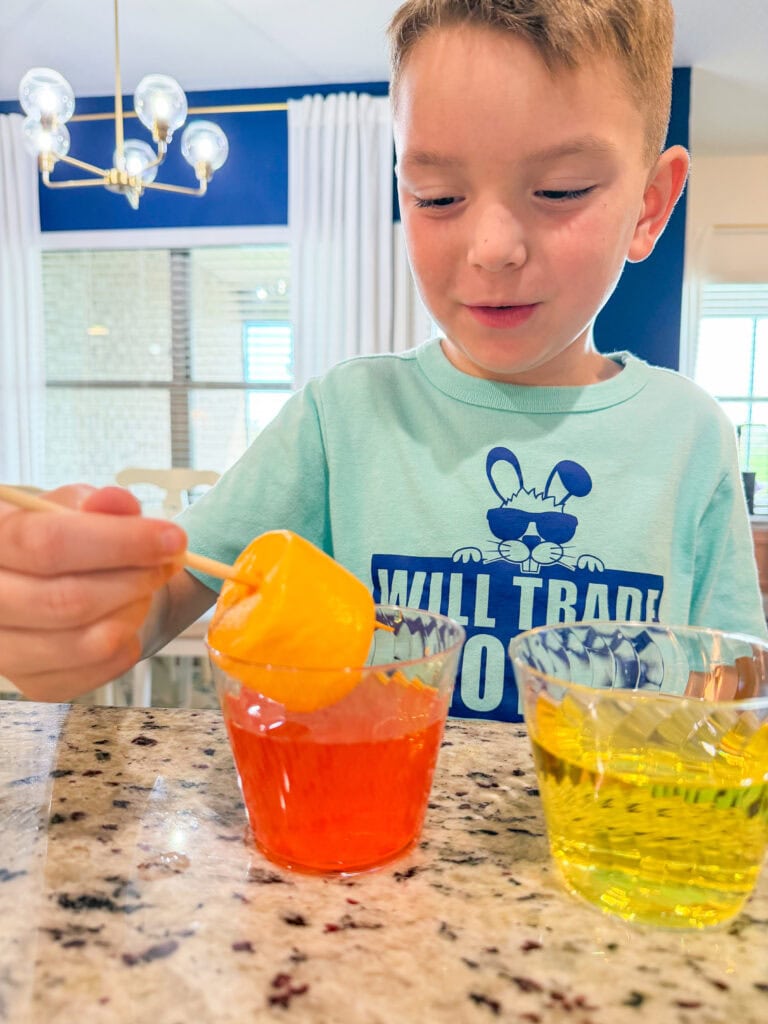 A young boy dips a yellow plastic egg on a stick into a cup of orange liquid, with another cup of yellow liquid beside him, on a kitchen counter. He wears a light blue shirt with a cartoon mouse.