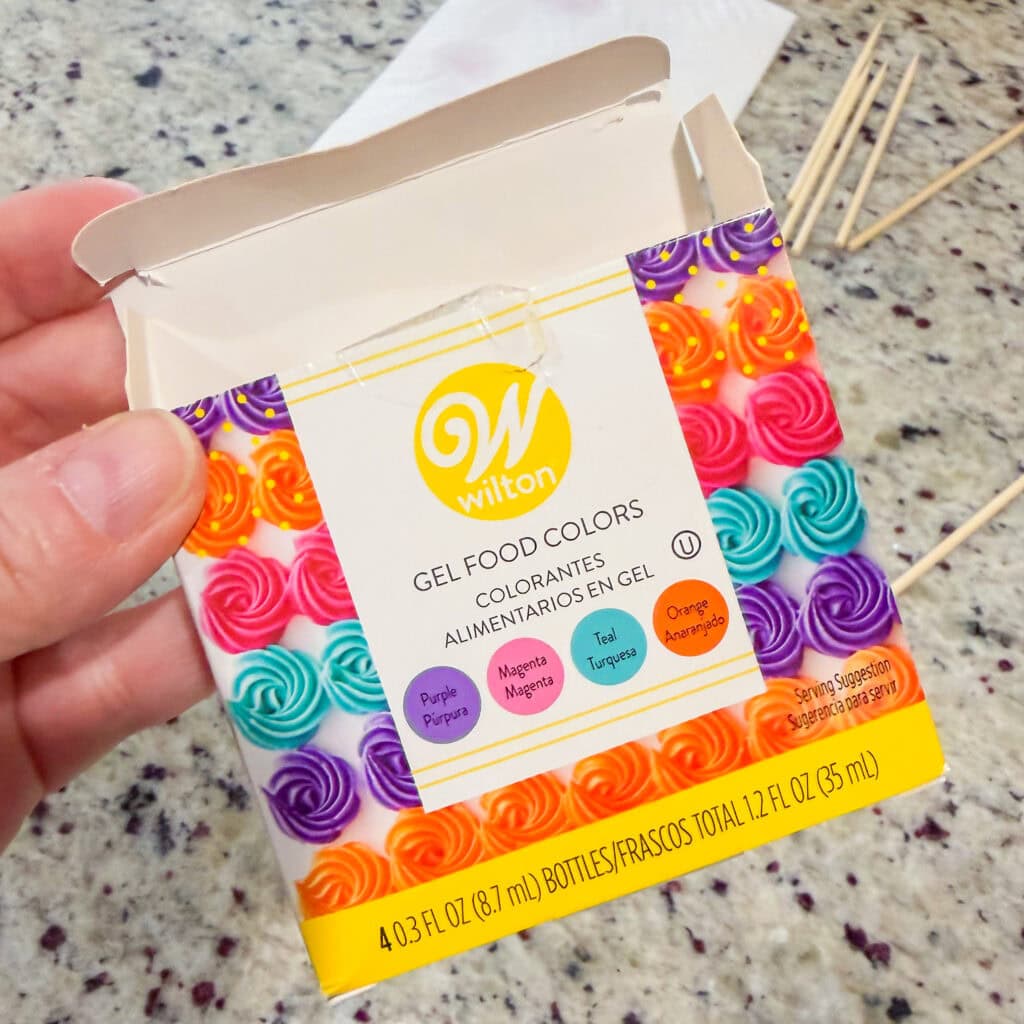 A hand holds an open box of Wilton gel food colors. The box features colorful icing swirls and lists four colors: Purple, Magenta, Teal, and Orange. Wooden sticks and a granite countertop are visible in the background.