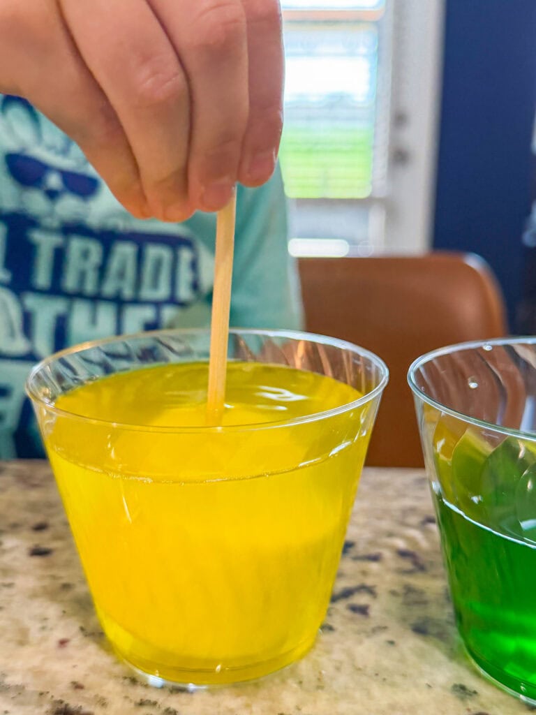 A hand stirs a clear plastic cup filled with bright yellow liquid using a wooden stick. Another cup with green liquid is nearby, both on a speckled countertop.