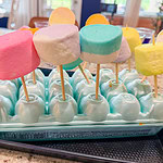 Large pastel-colored marshmallows on sticks are displayed upright in an empty blue egg carton on a kitchen counter. The egg carton helps hold the marshmallow pops in place.