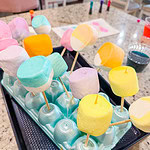 Colorful marshmallows on wooden skewers stand upright in an egg carton, set on a kitchen counter, with baking supplies and food coloring visible in the background.