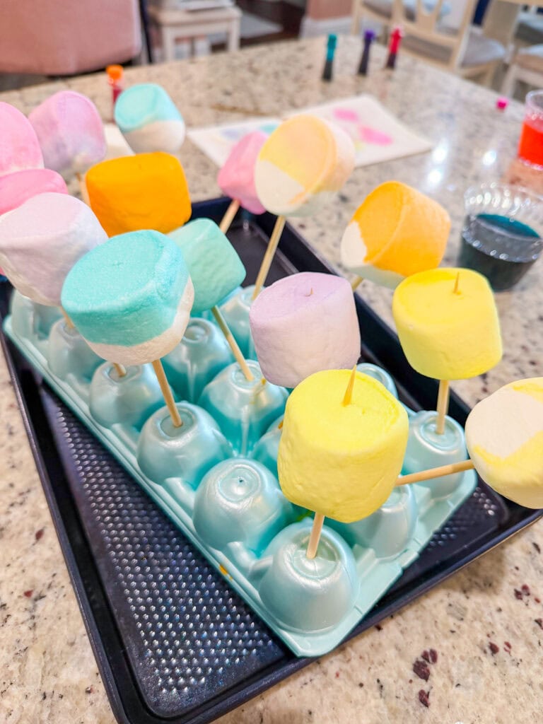 Colorful marshmallows on wooden skewers stand upright in an egg carton, set on a kitchen counter, with baking supplies and food coloring visible in the background.