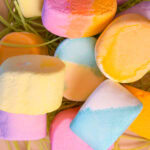 A close-up of colorful, pastel marshmallows in shades of yellow, pink, blue, and orange, piled together with green string candy underneath.