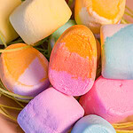 A close-up of colorful marshmallows in pastel shades of yellow, orange, pink, blue, and purple, arranged together with some green string-like candy in the background.