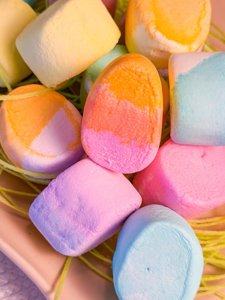 A close-up of colorful marshmallows in pastel shades of yellow, orange, pink, blue, and purple, arranged together with some green string-like candy in the background.