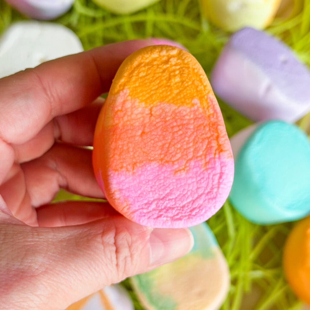 A hand holds a colorful, oval-shaped marshmallow with orange, yellow, and pink layers. More pastel marshmallows and green shredded paper are visible in the background.