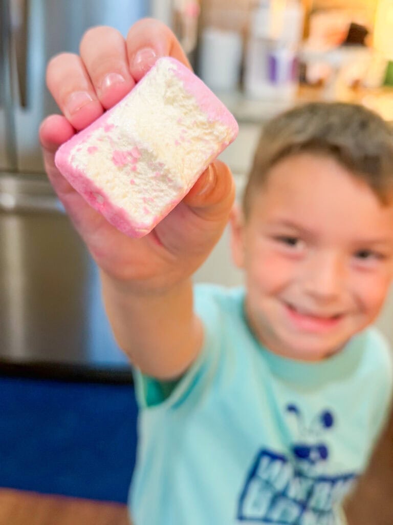 A smiling young boy holds up a pink and white square marshmallow toward the camera, with the focus on the marshmallow and a kitchen setting blurred in the background.