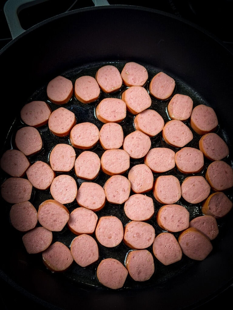 Sliced sausage pieces arranged in a single layer and cooking in a black skillet, with a bit of oil visible at the bottom of the pan.