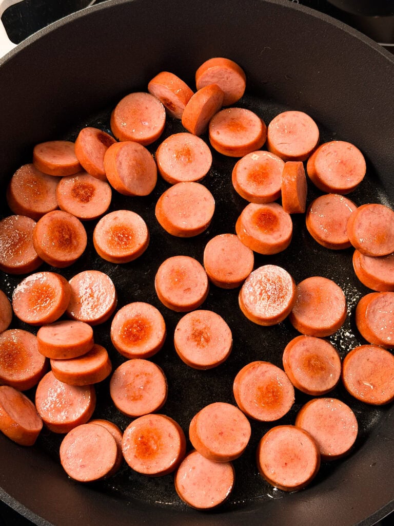 Sliced sausage rounds cooking in a black skillet, with some pieces slightly browned and sizzling in oil.