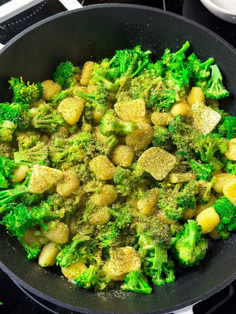 Broccoli florets and diced potatoes seasoned with spices are being cooked in a black frying pan on a stovetop.