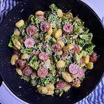 A black pan filled with gnocchi, sliced sausage, broccoli florets, and grated cheese, resting on a striped cloth with a wooden spoon nearby.