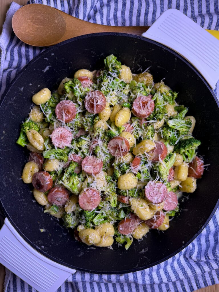 A black pan filled with gnocchi, sliced sausage, broccoli, and grated cheese, sits on a striped cloth with a wooden spoon nearby.