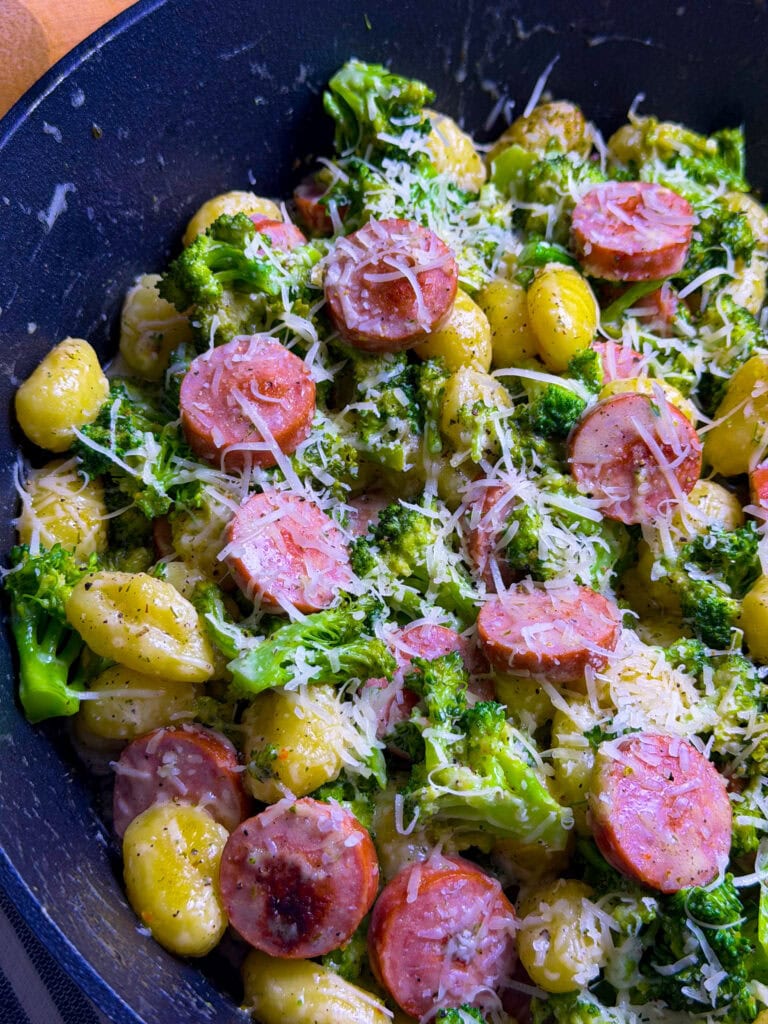 A skillet filled with gnocchi, sliced sausage, broccoli, and grated cheese, all mixed together and topped with black pepper.