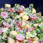 A close-up of creamy gnocchi mixed with broccoli, sliced sausage, and topped with grated parmesan cheese. The dish is in a large black skillet.
