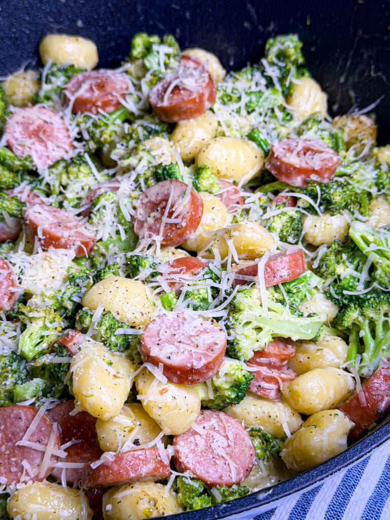 A close-up of creamy gnocchi mixed with broccoli, sliced sausage, and topped with grated parmesan cheese. The dish is in a large black skillet.