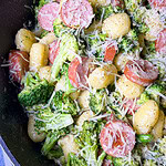 A close-up of a skillet filled with gnocchi, sliced sausage, broccoli florets, and shredded cheese, all coated in a creamy sauce.