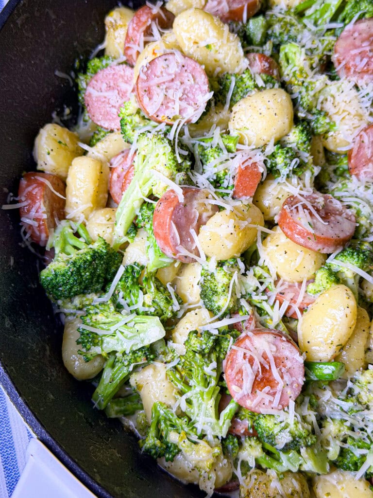 A close-up of a skillet filled with gnocchi, sliced sausage, broccoli florets, and shredded cheese, all coated in a creamy sauce.