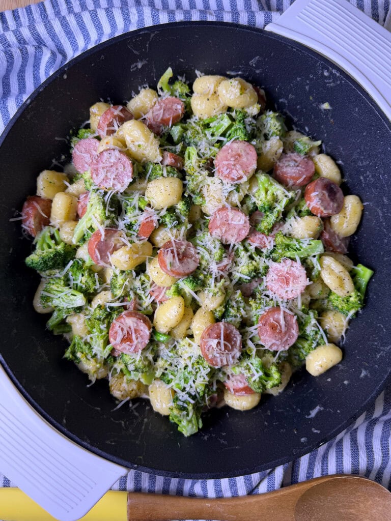 A skillet filled with gnocchi pasta, sliced sausage, and broccoli, all topped with grated cheese, sits on a striped cloth with a wooden spoon nearby.