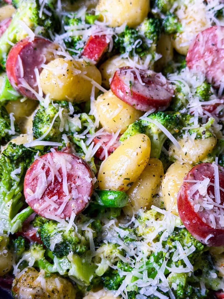 Close-up of a dish featuring gnocchi, sliced sausage, broccoli florets, and a generous sprinkle of shredded Parmesan cheese mixed with creamy sauce—a savory option to consider for your Easy Easter Treat Ideas.