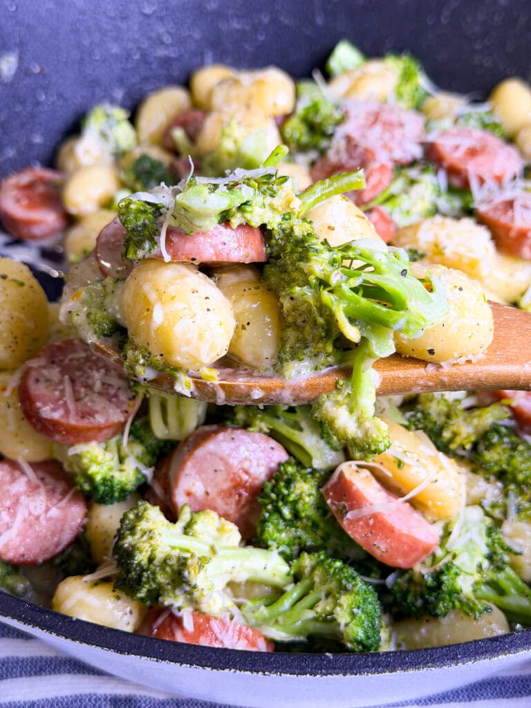 A close-up of a wooden spoon scooping gnocchi, broccoli, and sliced sausage in a creamy sauce topped with grated cheese. Served in a large black pan, this dish is one of those easy Easter treat ideas perfect for festive family dinners.