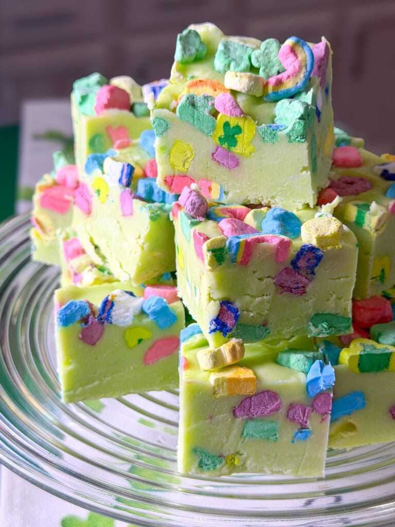 A stack of green fudge squares topped with colorful marshmallow shapes sits on a clear glass plate, making it one of the best Easy Easter Treat Ideas. The marshmallows resemble those from a popular breakfast cereal. The background is softly blurred.