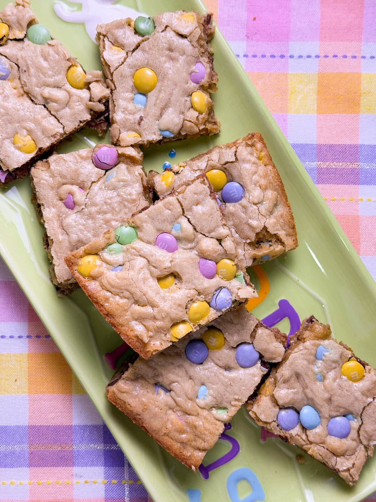 Seven blondie bars topped with pastel-colored candy-coated chocolates are arranged on a green rectangular plate. The plate sits on a pastel plaid tablecloth.
