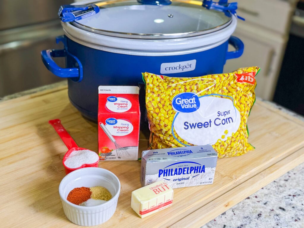 A blue Crockpot sits on a wooden counter with ingredients in front: a bag of sweet corn, cream cheese, unsalted butter, heavy whipping cream, a red measuring cup with flour, and four small bowls of spices.