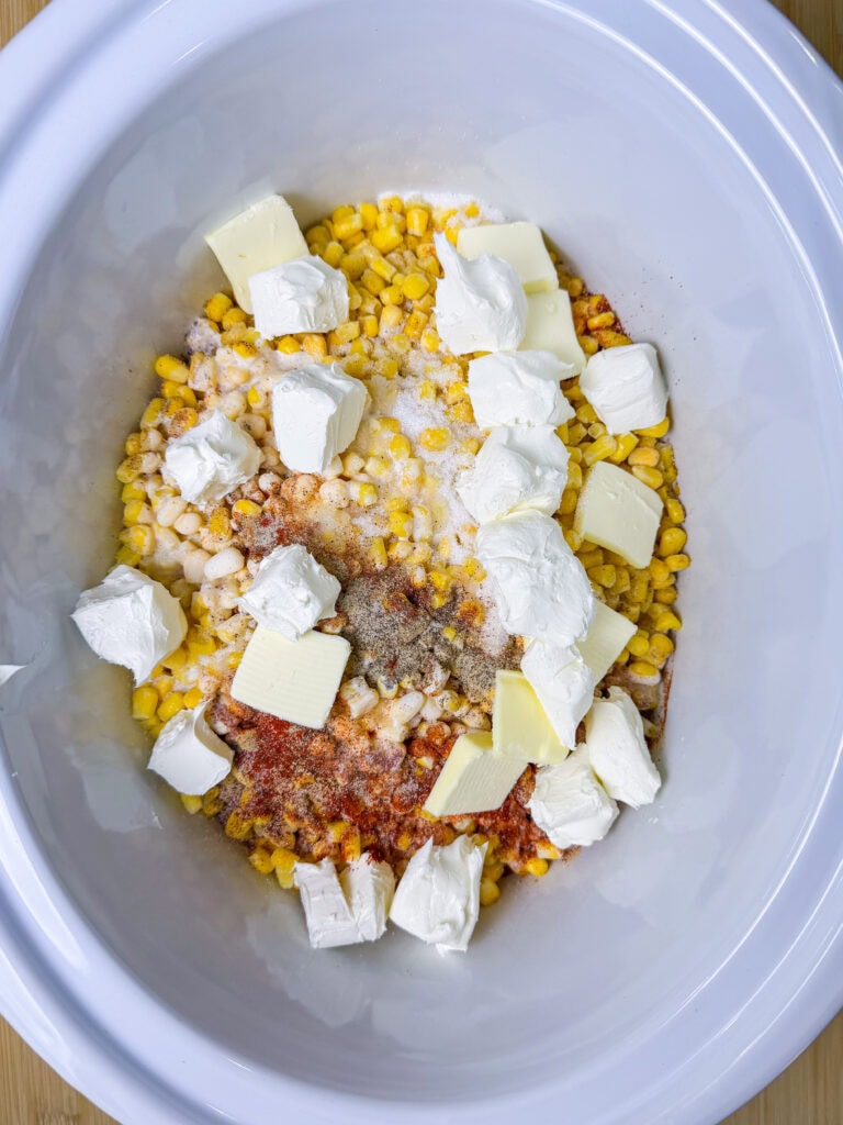 A white slow cooker filled with corn, chunks of cream cheese, butter, and various seasonings, ready to be mixed and cooked.