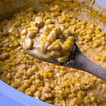 A wooden spoon holds creamy, seasoned corn kernels above a pot filled with more of the same creamy corn mixture.