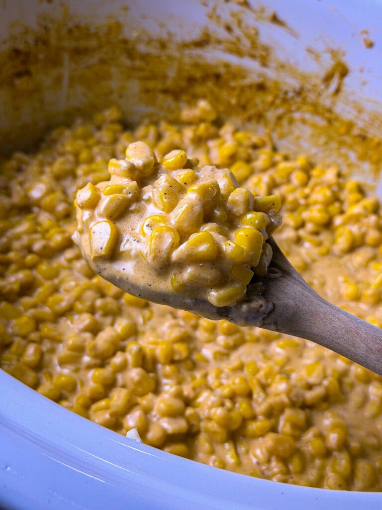A wooden spoon holds creamy, seasoned corn kernels above a pot filled with more of the same creamy corn mixture.
