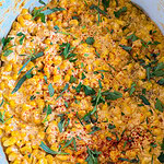 Creamy corn dish topped with chopped fresh herbs and a sprinkle of paprika, served in a blue bowl. The mixture appears rich and seasoned, with visible kernels and green garnish.