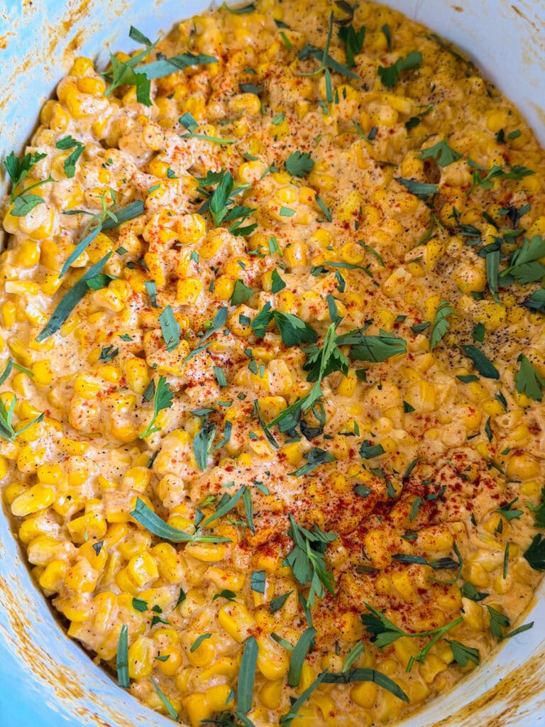Creamy corn dish topped with chopped fresh herbs and a sprinkle of paprika, served in a blue bowl. The mixture appears rich and seasoned, with visible kernels and green garnish.