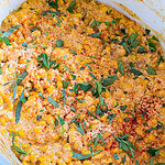 Creamy corn casserole topped with fresh chopped herbs and a sprinkle of paprika, served in a white dish. The casserole has a golden, cheesy appearance with visible corn kernels and herbs.