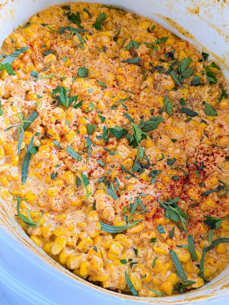 Creamy corn casserole topped with fresh chopped herbs and a sprinkle of paprika, served in a white dish. The casserole has a golden, cheesy appearance with visible corn kernels and herbs.