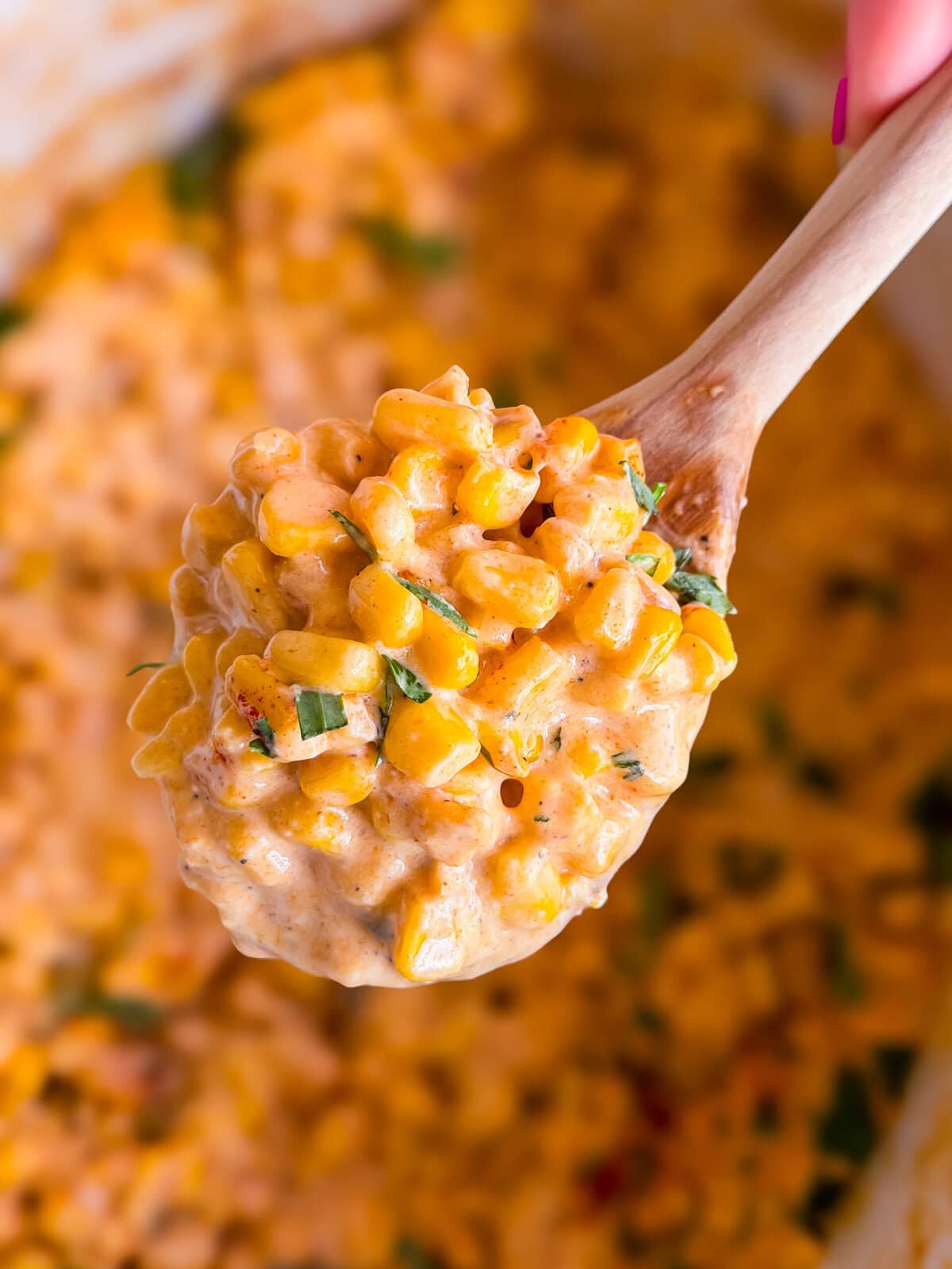 A wooden spoon holds a scoop of creamy corn mixed with herbs, with more of the creamy corn mixture blurred in the background.