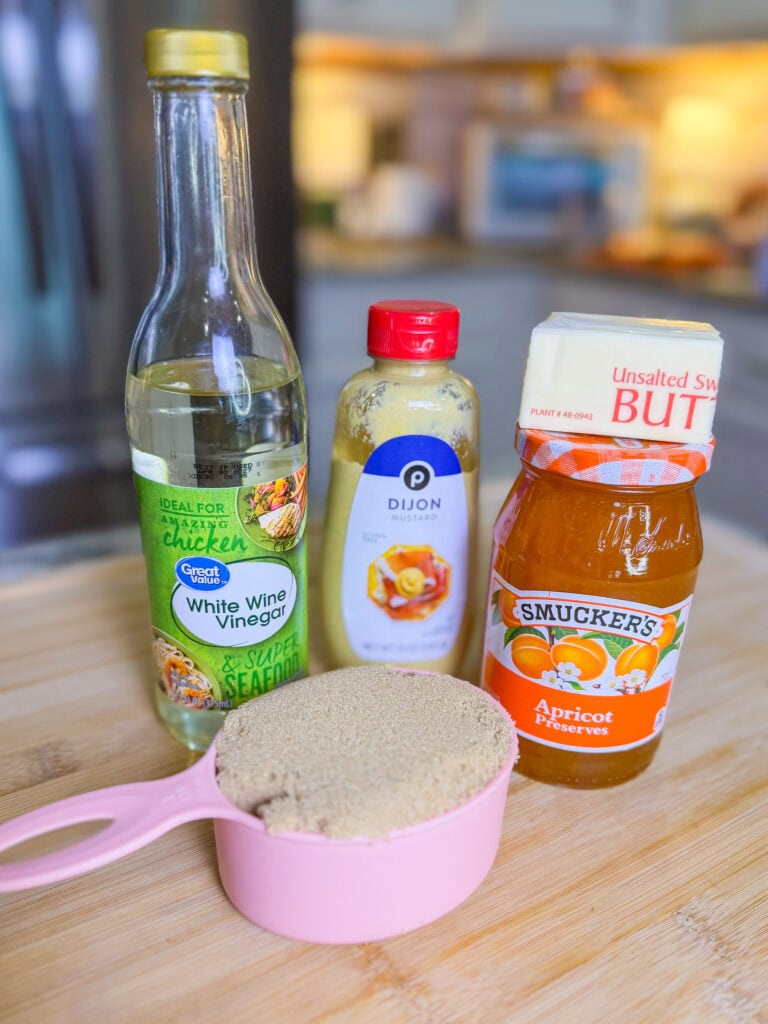 A bottle of white wine vinegar, a bottle of Dijon mustard, a jar of apricot preserves, and a pink measuring cup filled with brown sugar are arranged on a wooden surface in a kitchen.