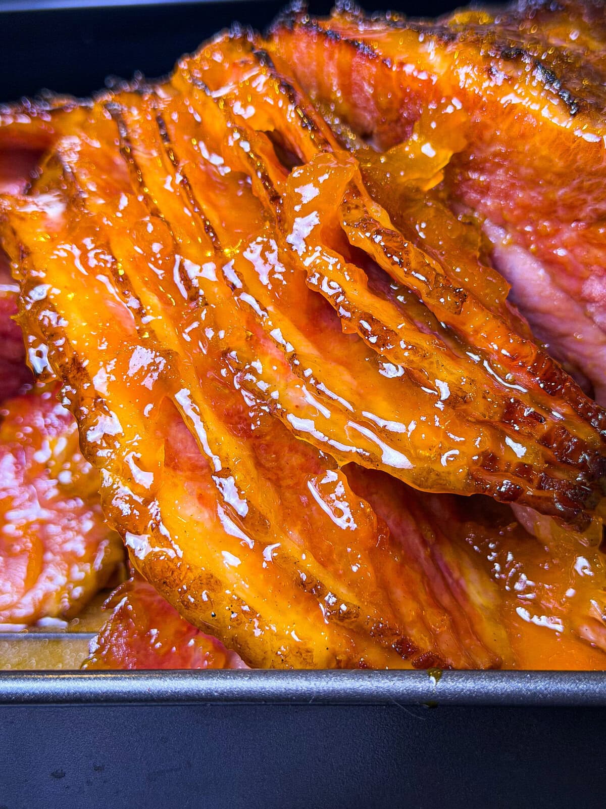 Close-up of sliced glazed ham, with a shiny, caramelized coating, stacked in a baking pan. The ham appears juicy and tender with crispy edges.