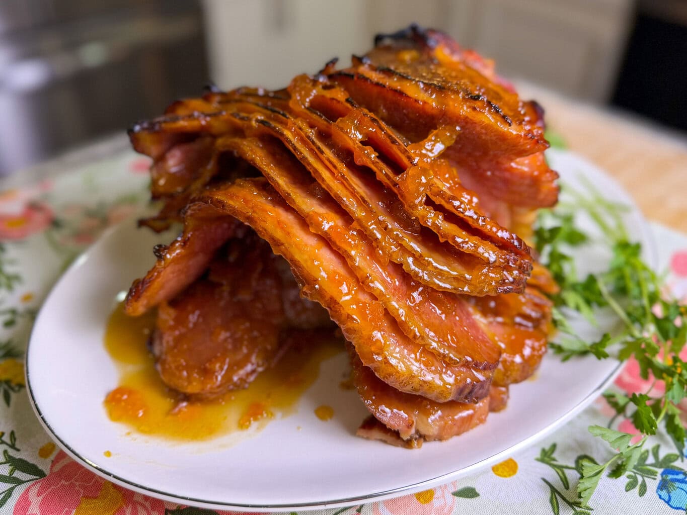 A plate of glazed, sliced ham arranged in layers, covered in a shiny, caramelized sauce, sits on a floral tablecloth with fresh herbs in the background.