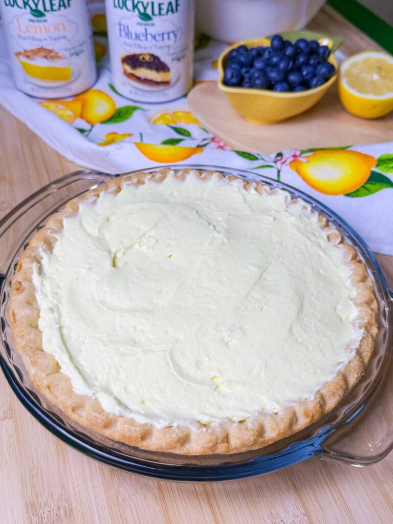 A lemon cream pie with a golden crust sits in a glass pie dish. Behind it are cans of pie filling, a bowl of fresh blueberries, half a lemon, and a lemon-patterned cloth on a wooden surface.