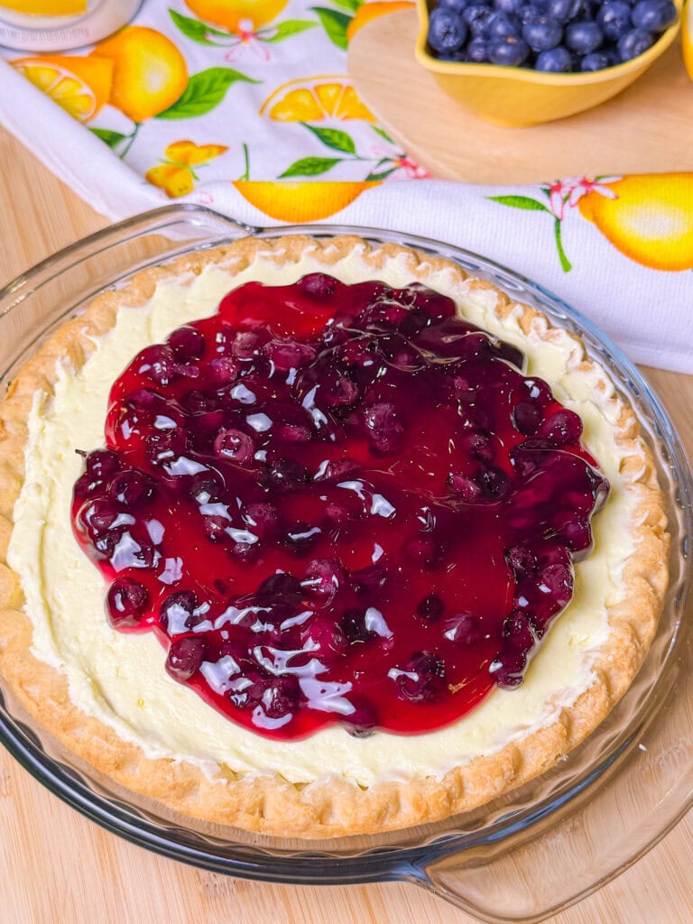 A pie dish filled with a cheesecake topped with a glossy blueberry sauce sits on a wooden surface, with a bowl of fresh blueberries and a lemon-patterned cloth in the background.