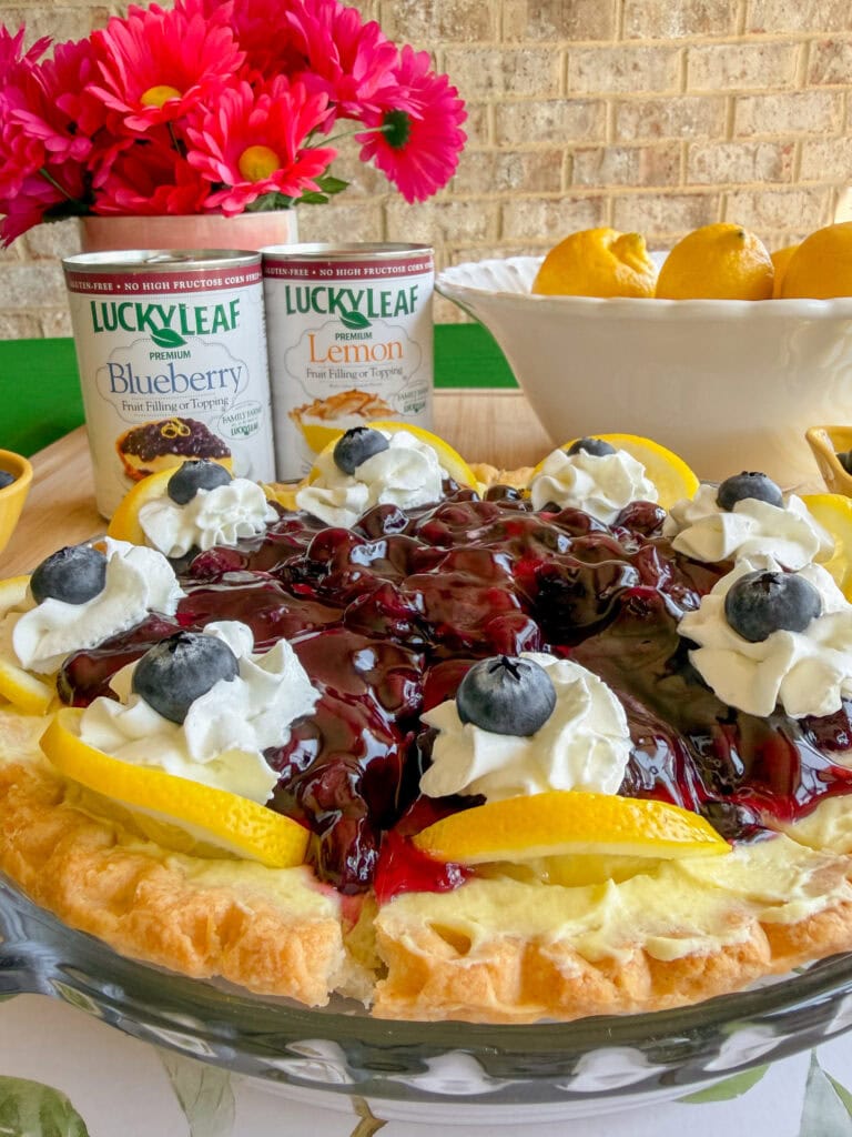 A blueberry lemon pie topped with whipped cream, blueberries, and lemon slices. In the background are cans of Lucky Leaf blueberry and lemon pie filling, a bowl of lemons, and a vase of pink flowers.