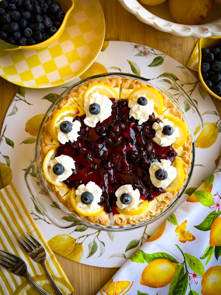 A blueberry lemon pie topped with whipped cream, lemon slices, and blueberries sits on a lemon-patterned placemat, surrounded by bowls of blueberries, a yellow-checked napkin, and silver forks.