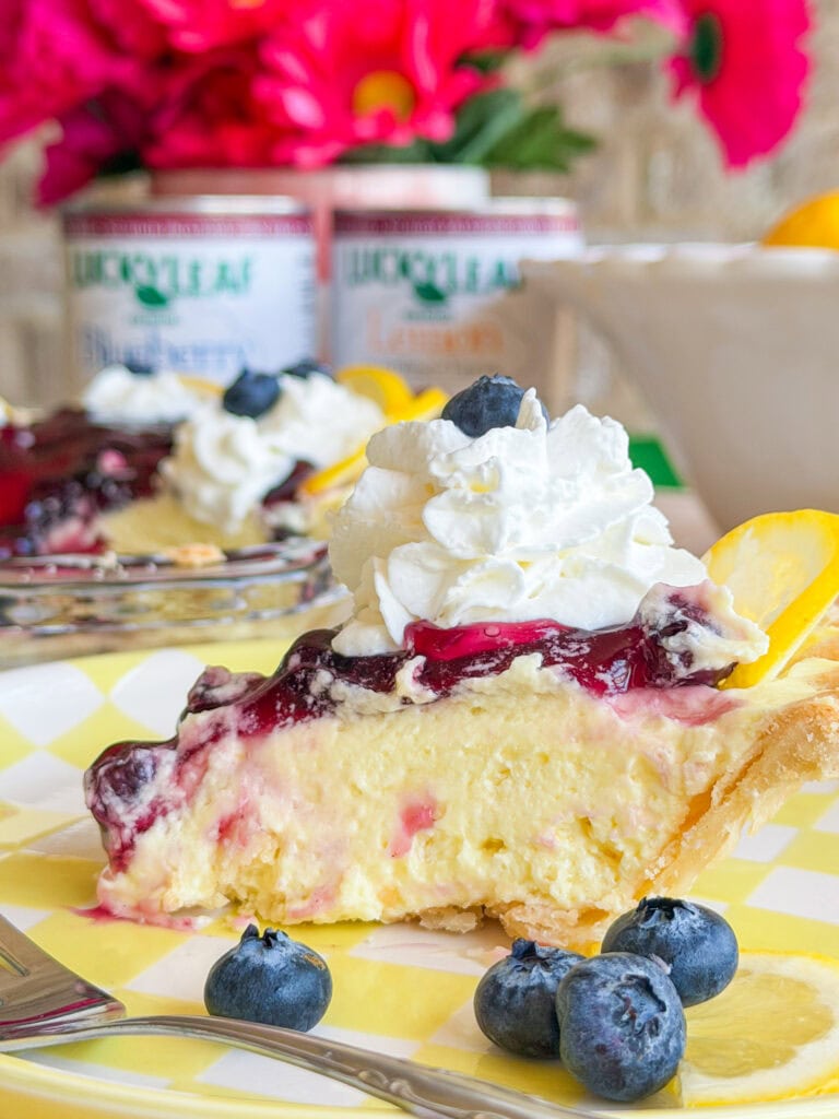 A slice of creamy pie topped with blueberry sauce, whipped cream, and a lemon slice sits on a yellow checkered plate, garnished with fresh blueberries. Bright flowers and canned fruit are blurred in the background.