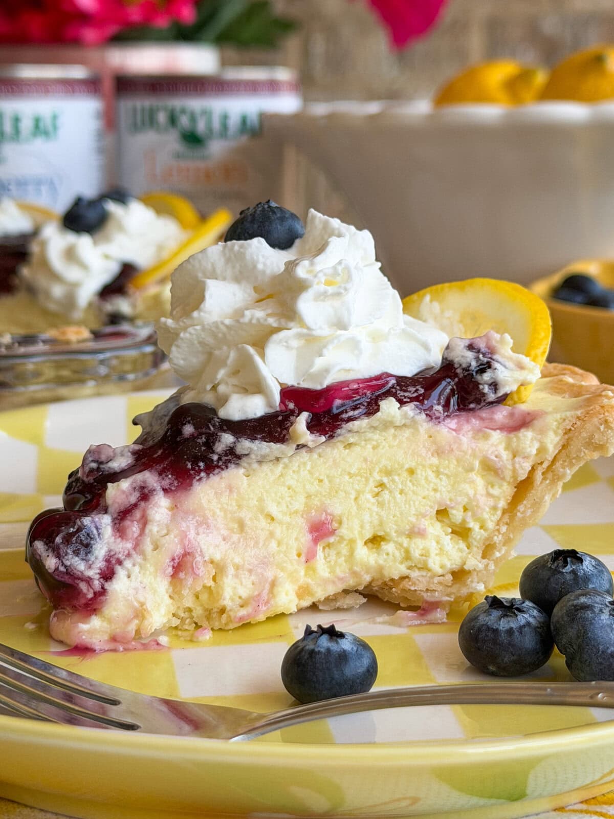 A slice of creamy pie topped with whipped cream, blueberry sauce, and a blueberry, served on a yellow plaid plate with fresh blueberries and a lemon slice, with a fork beside it.