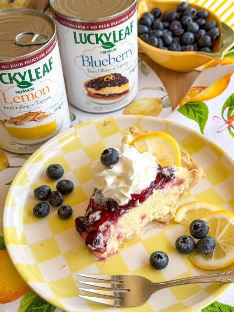 A slice of cheesecake topped with blueberry sauce, whipped cream, a lemon slice, and blueberries sits on a yellow checkered plate. Nearby are cans of Lucky Leaf lemon and blueberry filling, a bowl of blueberries, and lemon slices.