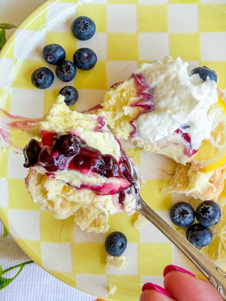 A plate with a slice of dessert topped with whipped cream and blueberry sauce, surrounded by fresh blueberries. A hand holding a fork is cutting a bite from the dessert. The plate has a yellow and white checkered pattern.