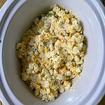 A white slow cooker filled with a creamy potato mixture that includes shredded cheese and small pieces of vegetables, sitting on a wooden surface.