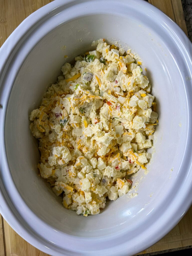 A white slow cooker filled with a creamy potato mixture that includes shredded cheese and small pieces of vegetables, sitting on a wooden surface.