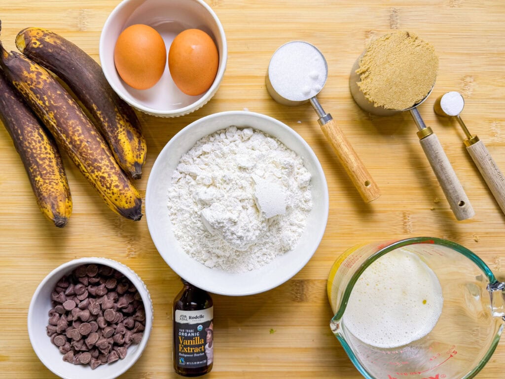 Overhead view of baking ingredients on a wooden surface: ripe bananas, eggs, flour, granulated sugar, brown sugar, chocolate chips, vanilla extract, and a measuring cup with foamy liquid, likely milk.
